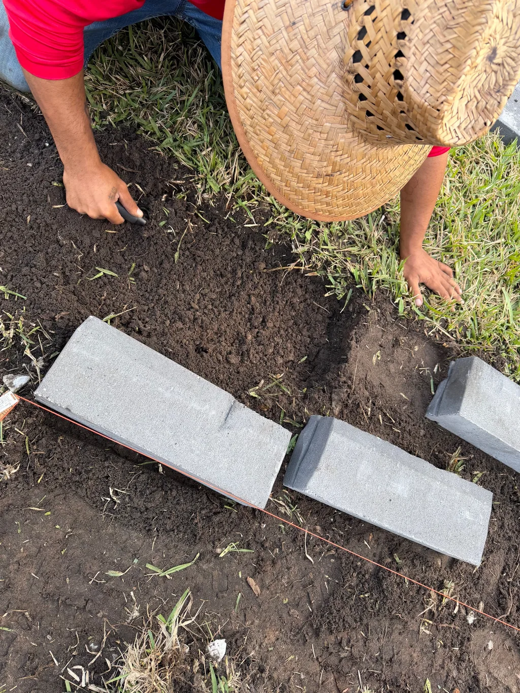 Hands setting concrete edging stones into a shallow soil trench beside a cut lawn